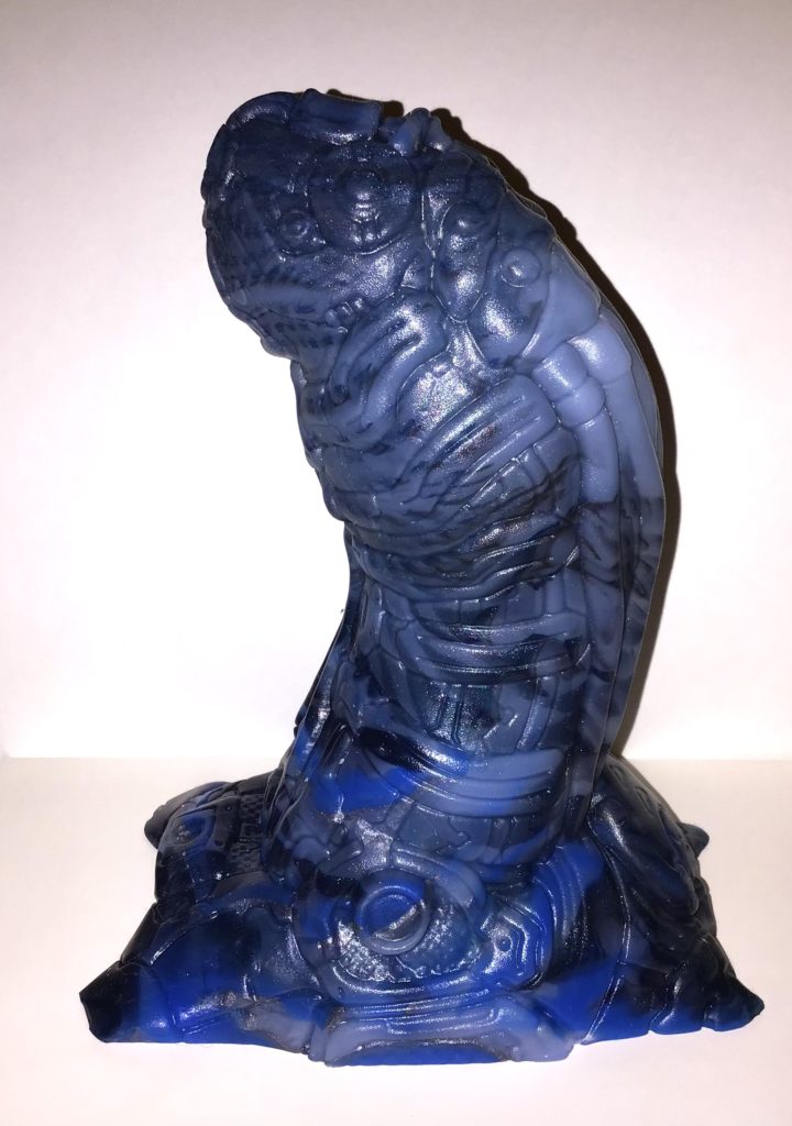 A stout, mechanical, humanoid penis with a strong curve. It is highly detailed. The pour is a marble of blue, black, and foggy grayish silicone.
