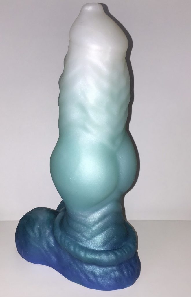 The top half of the shaft features subtle ridged texture. The bottom half of the shaft features a knot, followed by a textured stem. The base is a furry sheath with balls. It's a fade from white at the top to light teal blue in the middle and dark teal blue at the base.