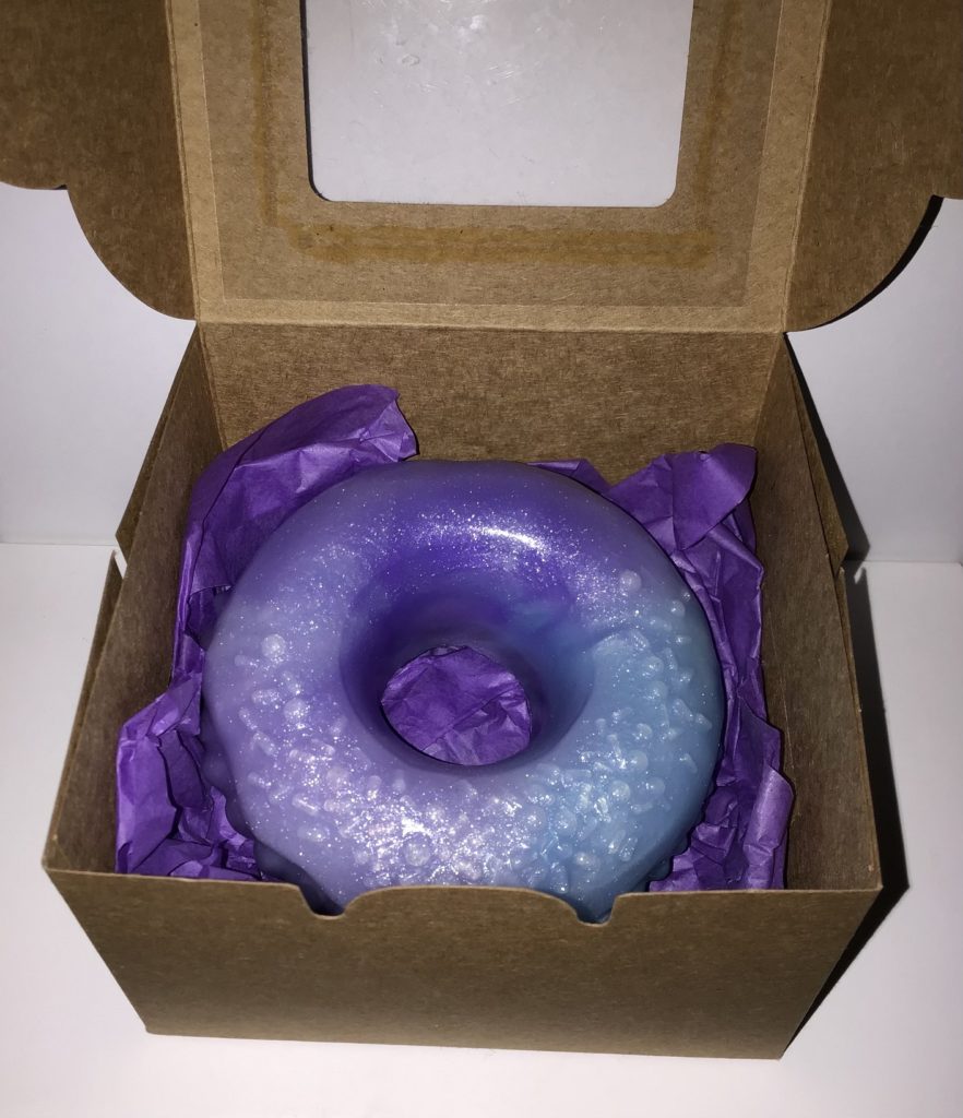 Silicone donut with sprinkles on top inside a small cardboard box. It's light blue and purple with a shimmery white highlight.