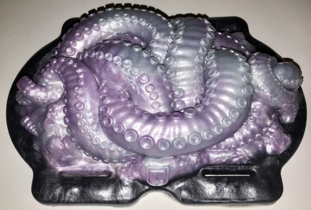 A mass of writhing, hihgly textured tentacles atop a base through which straps may be threaded. The base is black and the tentacles are pink and silver.